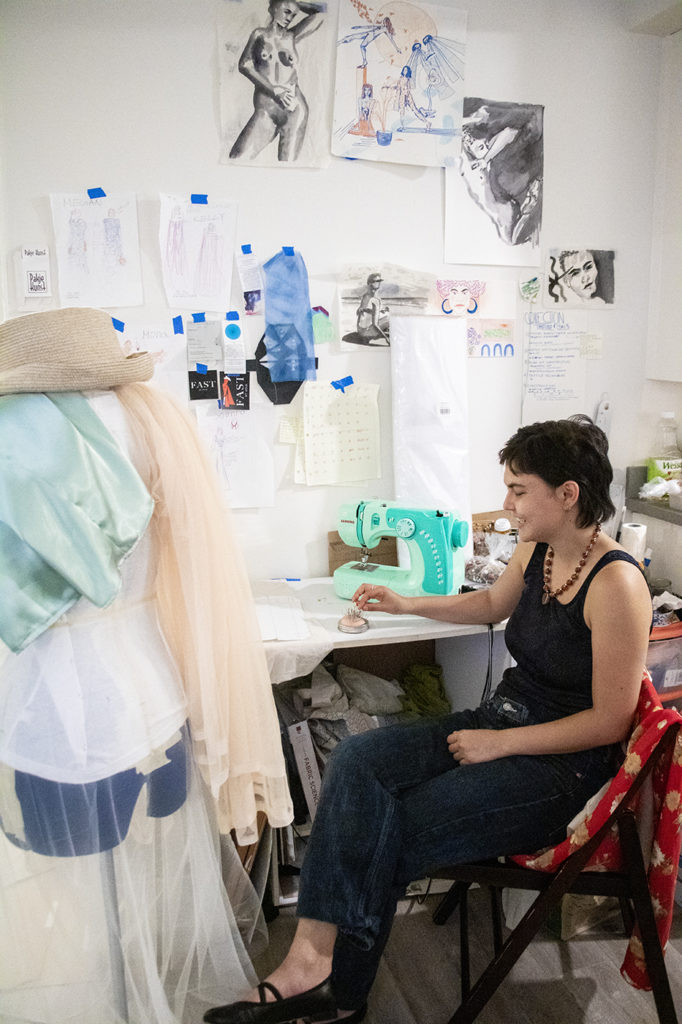 Lin sits at a desk, framed by a mannequin draped with fabrics on the left and notes taped to the wall above. For Lin, the COVID-19 pandemic resulted in a more limited access to materials, leading to more avant-garde creations. (Finn Chitwood/Daily Bruin)
