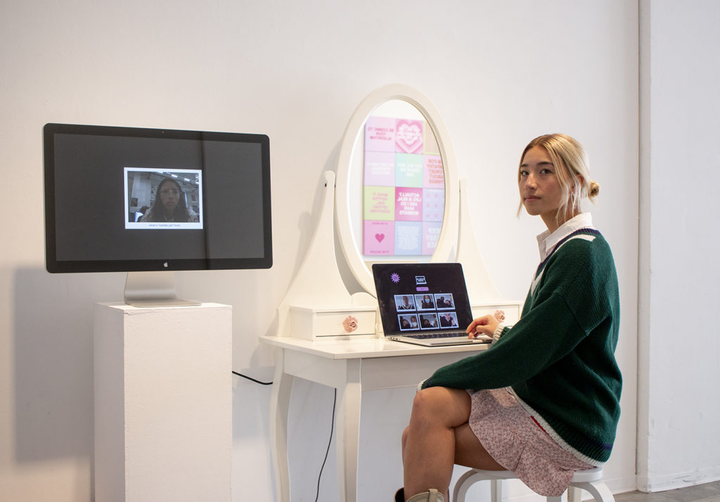 Man sits at a vanity in front of a laptop displaying snapshots, which are also seen on the computer beside the vanity. For "Secrets From a Girl," Man said she wanted to transform the exhibit space into one that looked similar to her childhood bedroom. (Christine Kao/Daily Bruin staff)