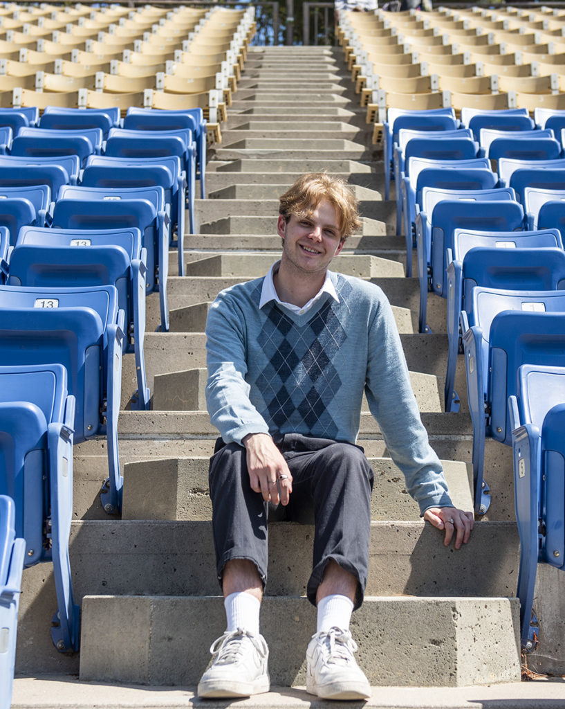 Drinker sits on steps between rows of blue bleachers. With vulnerable lyrics based on personal experiences, Drinker said he aims for listeners to find hope in the stories his songs tell. (August Suchecki/Daily Bruin)