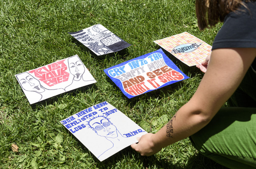 Five multicolored prints lie spread out atop grass. With pieces featuring phrases like "Trust Your Eyes," Markham has found ways to blend her artisits and musical endeavors whether that be through adding lyrics to her pieces or through creating graphic scores. (August Suchecki/Daily Bruin)
