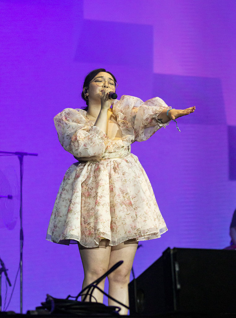 In front of a purple-lit screen, mxmtoon reachs to the crowd, wearing a floral dress. The singer-songwriter frequently interacted with audience members throughout her evening set. (Megan Cai/Assistant Photo editor)