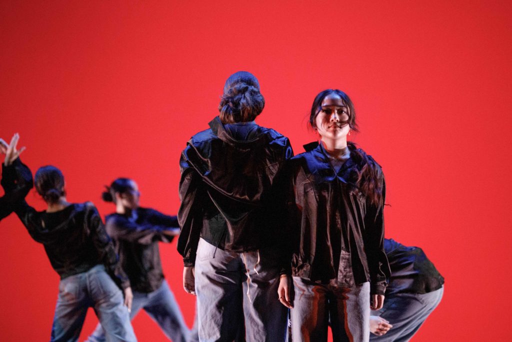 Performers dressed in black stand side by side in front of other dancers in motion and a red backdrop. The annual student-led dance and art showcase will be hold performances Friday and Saturday in Kaufman Hall. (Grace Wilson/Daily Bruin)