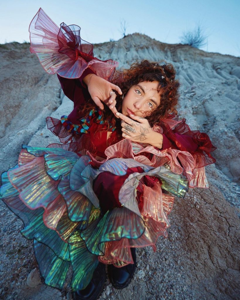 Dressed in a shimmery multicolored dress, Indigo DeSouza sits on rocky ground. The indie-alternative artist is set to release her third album "All of This Will End" Friday. (Courtesy of Angella Choe)