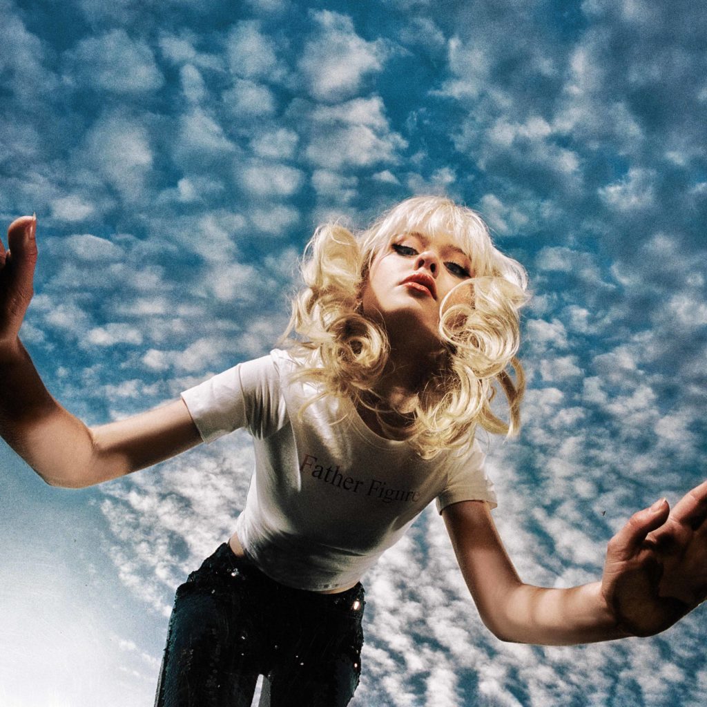 With a blue and cloud-filled sky behind her, Maisie Peters bends down towards the camera on the cover of her sophomore album “The Good Witch,” which is set to release June 16. Courtesy of Gingerbread Man Records)