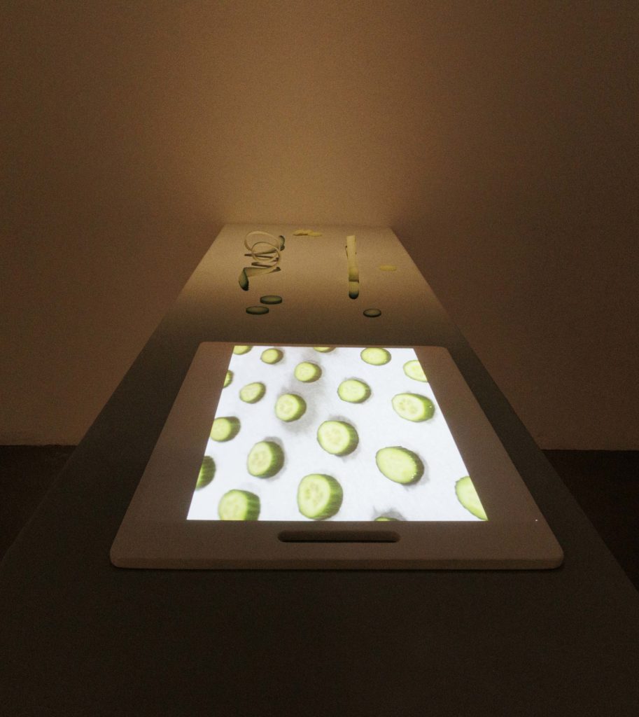 Cucumbers sliced into irregular shapes and spirals lie on a table next to a cutting board with an image of round slices of cucumbers projected onto it. When it comes to her art, Choi said she gravitates towards items that are often overlooked. (Grace Wilson/Daily Bruin)