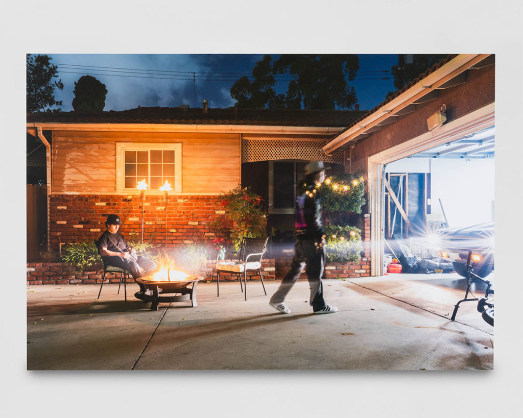 Taken in the driveway of a suburban home, the photograph depicts two lawnchairs by a fire pit, where one boy sits while another walks away. Christina Fernandez said her piece, "Suburban Nightscapes #1 (Max and Diego)," uses motion blur to illustrate the progression of time. (Photo by Yubo Dong, Courtesy of Charlie James Gallery)