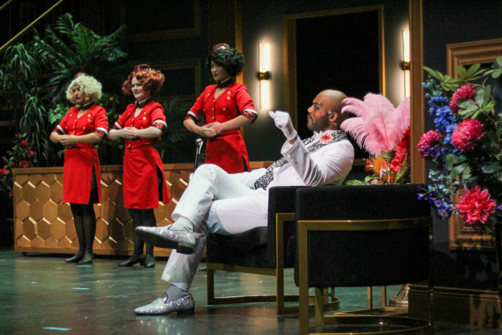 Dressed in a silver and white suit, LaMarcus Miller's Mr. Lucian sits onstage surrounded by guest services women. Exploring themes of desire and disease, the opera performances are split between two casts. (Courtesy of Robert Baker)