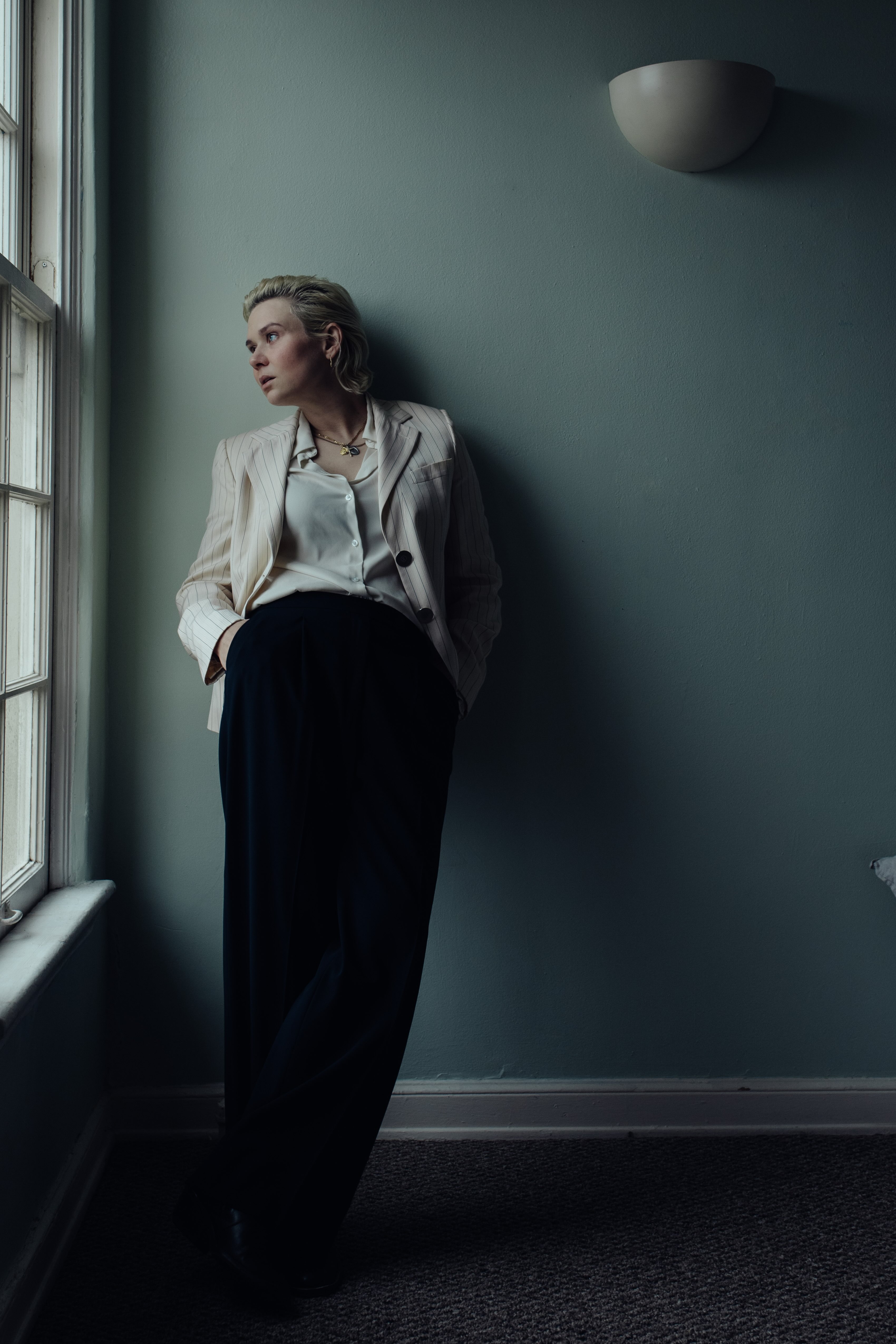 Amy Kuney stands against a pale blueish-gray wall while looking out of a window and wearing a white suit jacket and black pants. Kuney, who is originally from Oklahoma, releases her own music as Ames while co-writing songs for stars such as Kelly Clarkson and Kali Uchis. (Courtesy of Alexandra Petruck)