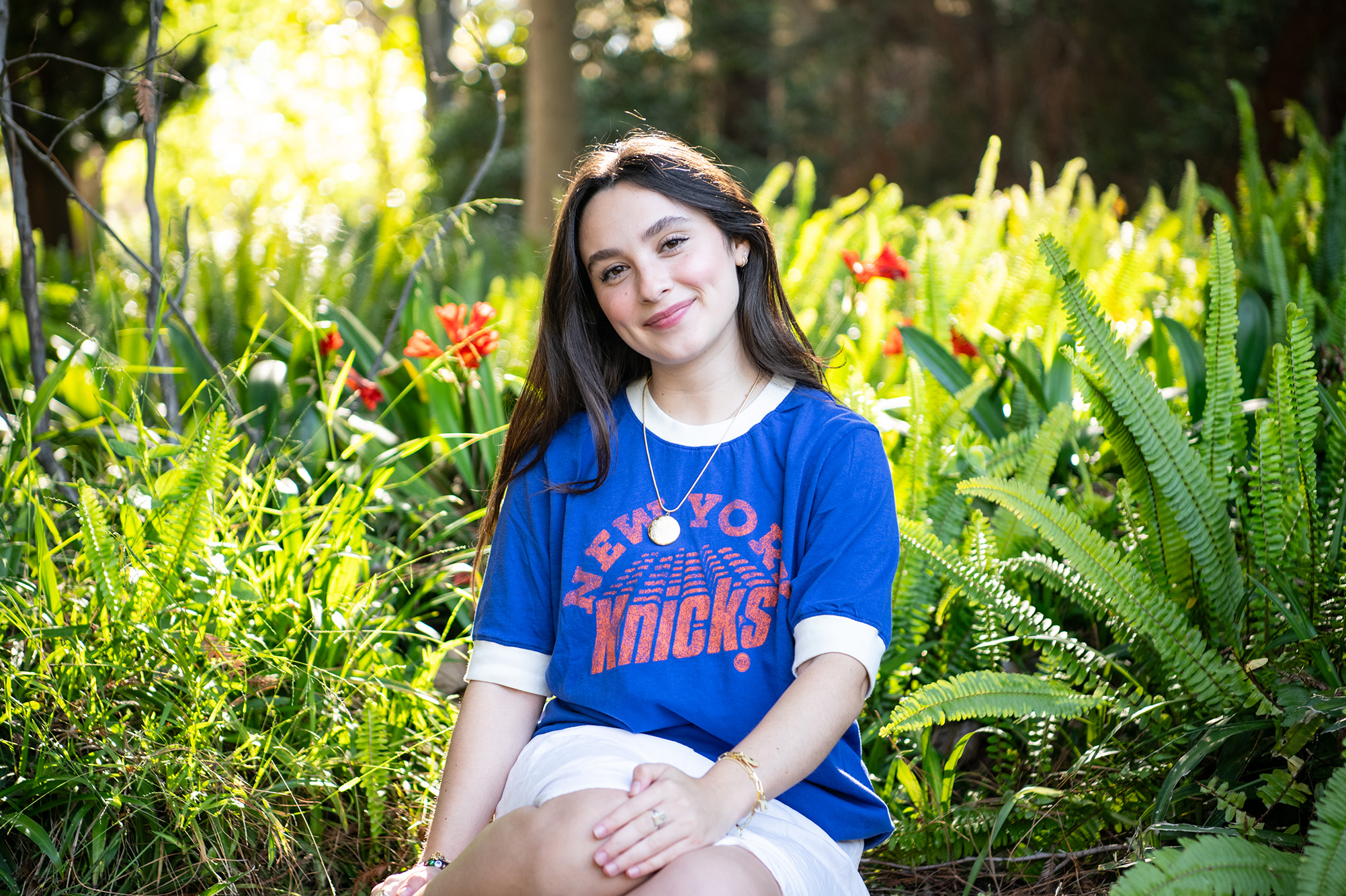 Ava Ulloa wears a blue New York Knicks shirt and sits surrounded by plants, including red flowers and ferns. Ulloa has taken on private lessons with adjunct professor Michele Weir, who teaches in the UCLA global jazz studies department, to better learn the genre of jazz. (Amanda Velasco/Daily Bruin)