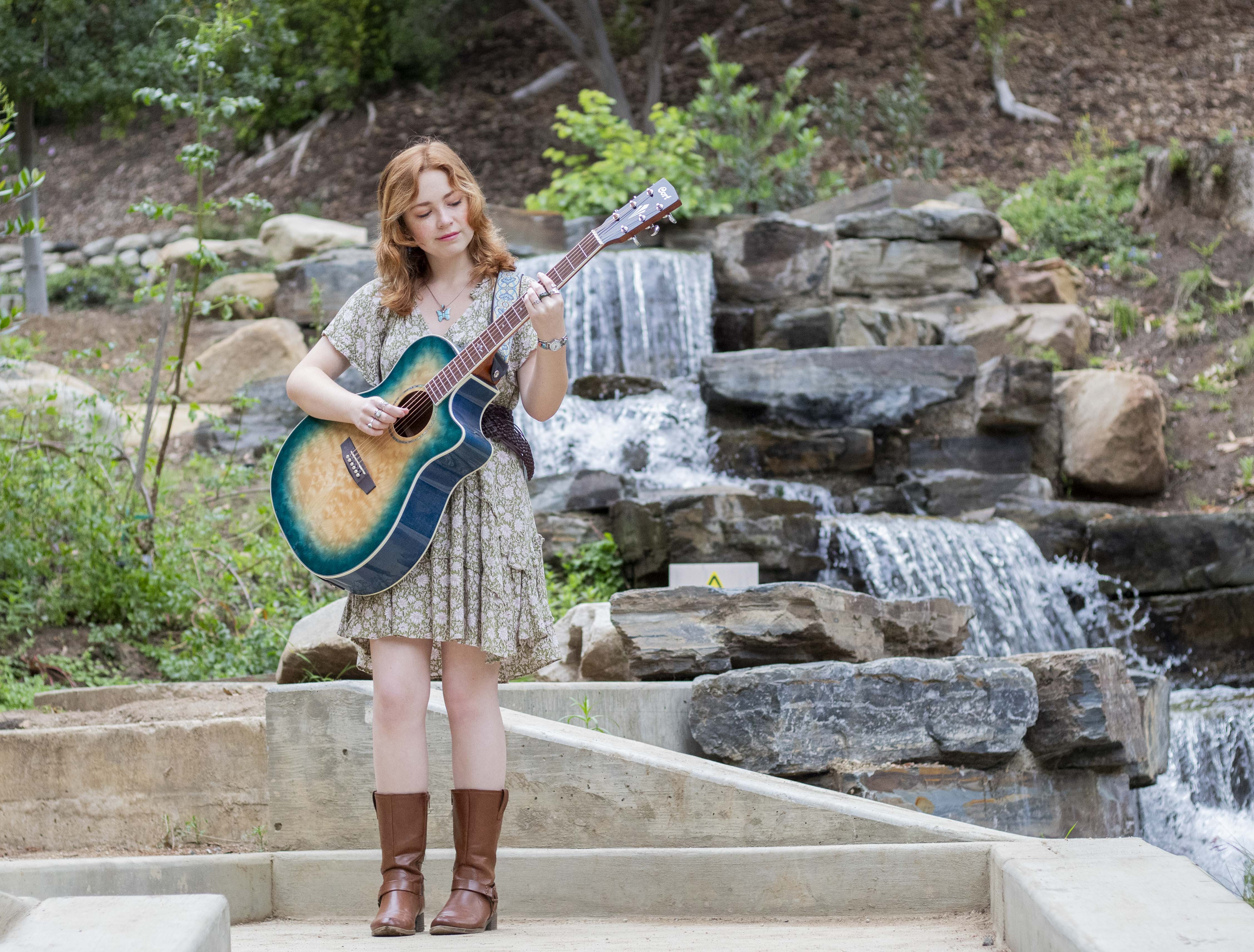 Ella Gibson stands next to a small waterfall with her guitar while wearing a green dress and brown leather boots. The second-year music industry student will perform her unreleased song "Patch" at Spring Sing on Friday. (Danielle Anz/Daily Bruin senior staff)