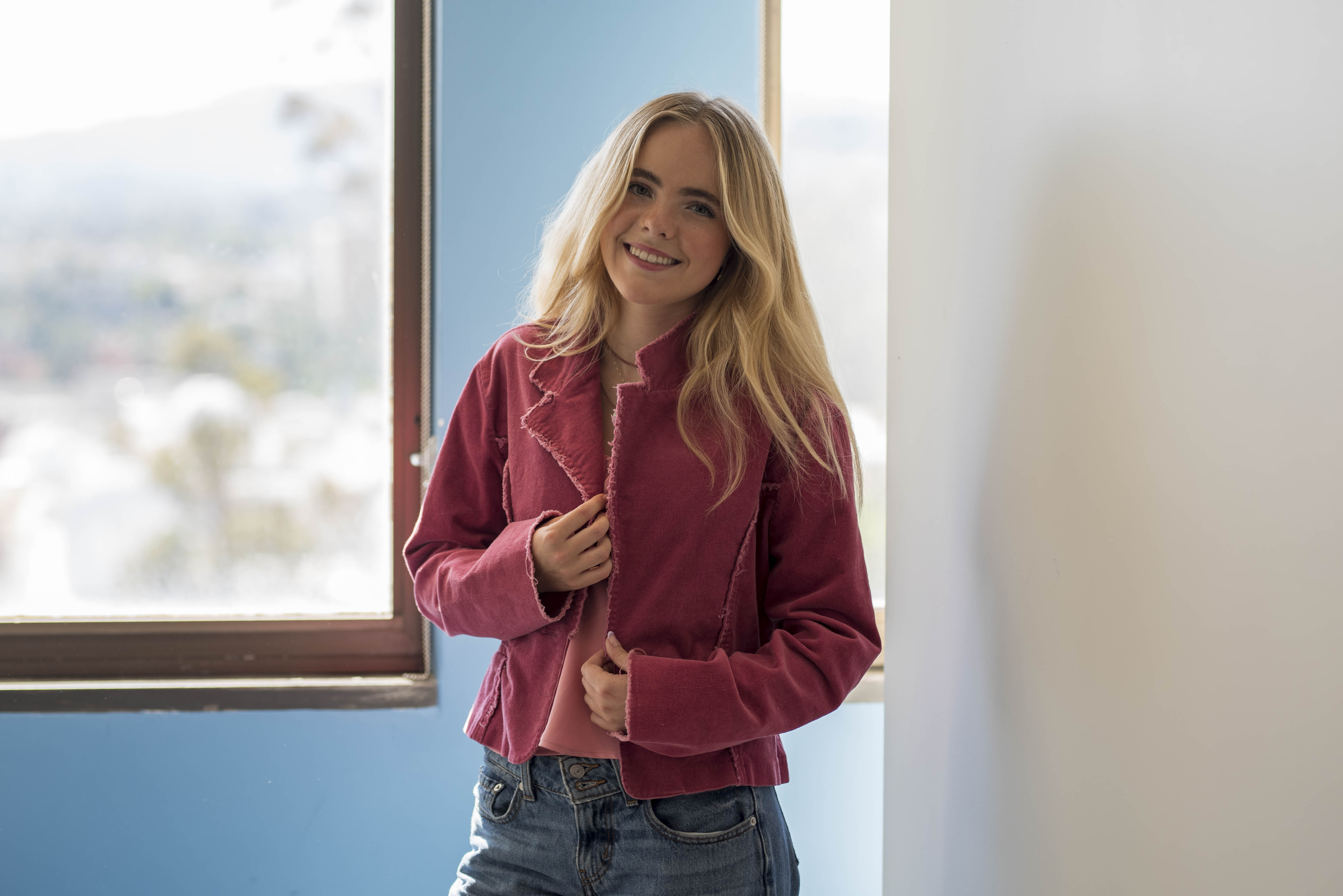 Chappell stands in front of a blue wall and window while wearing a pink jacket and denim jeans. The student musician said she enjoys building community through her DJ sets. (Brianna Carlson/Daily Bruin staff)