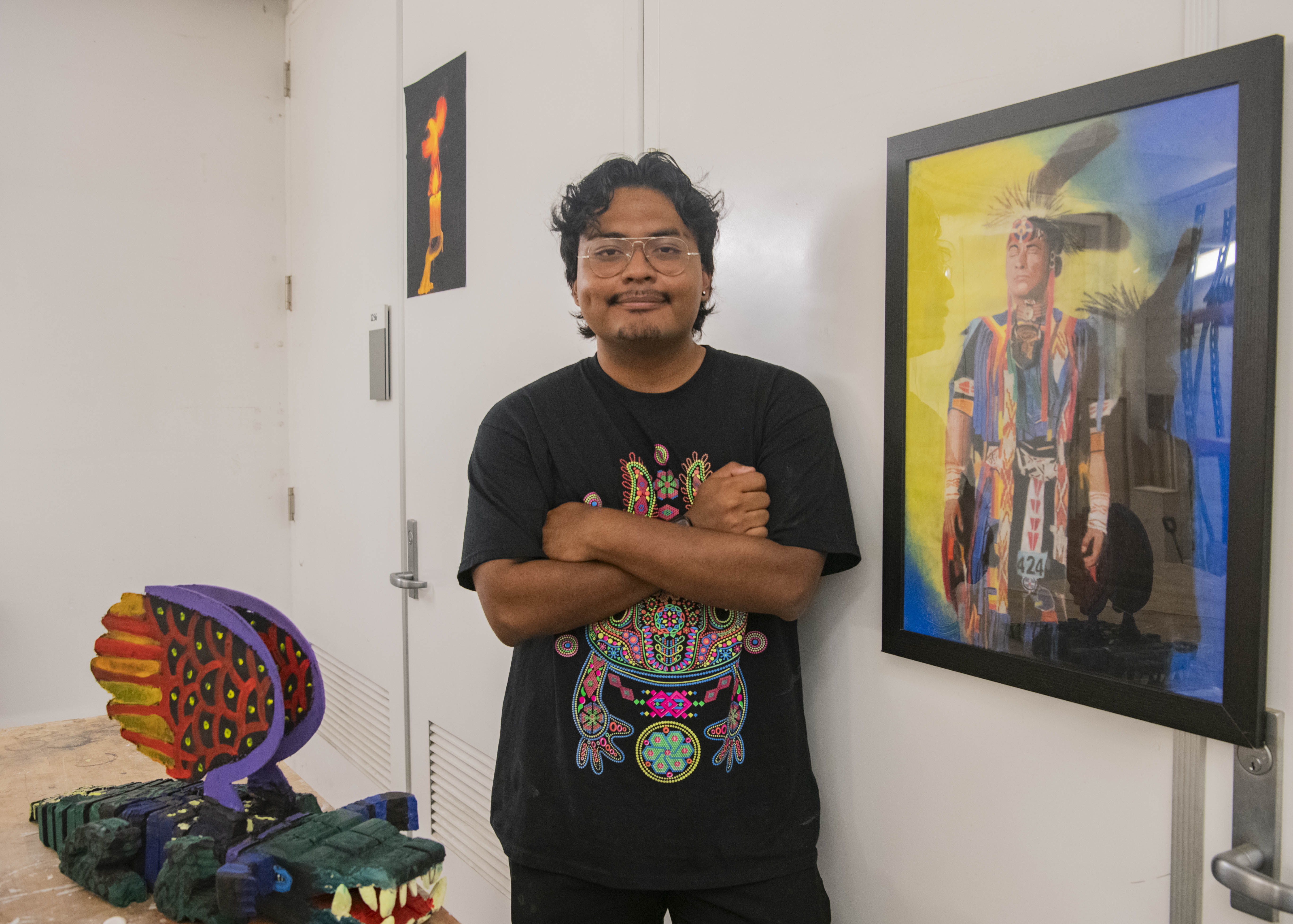 Ramirez stands in between two art pieces with his arms crossed over his chest. The multimedia artist said he began drawing at a very young age due to his love for comic books, often creating portraits of those around him. (Isabella Appell/Daily Bruin)