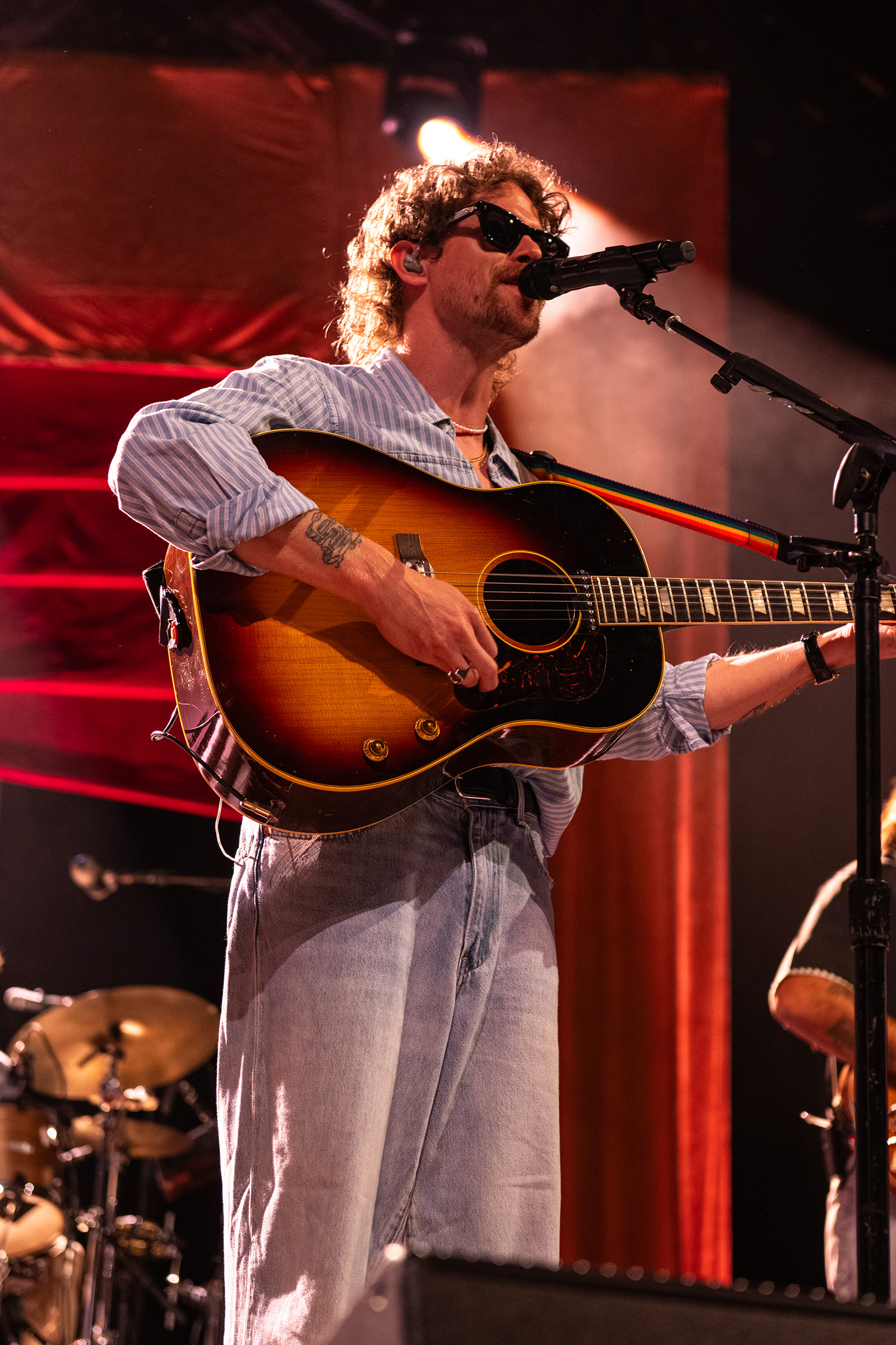 Caamp's lead vocalist wears a button-up shirt and sunglasses while singing and playing an acoustic guitar. Taylor Meier was often spotlighted on the venue's screens while performing in a relaxed and comfortable manner. (Isabella Appell/Daily Bruin)