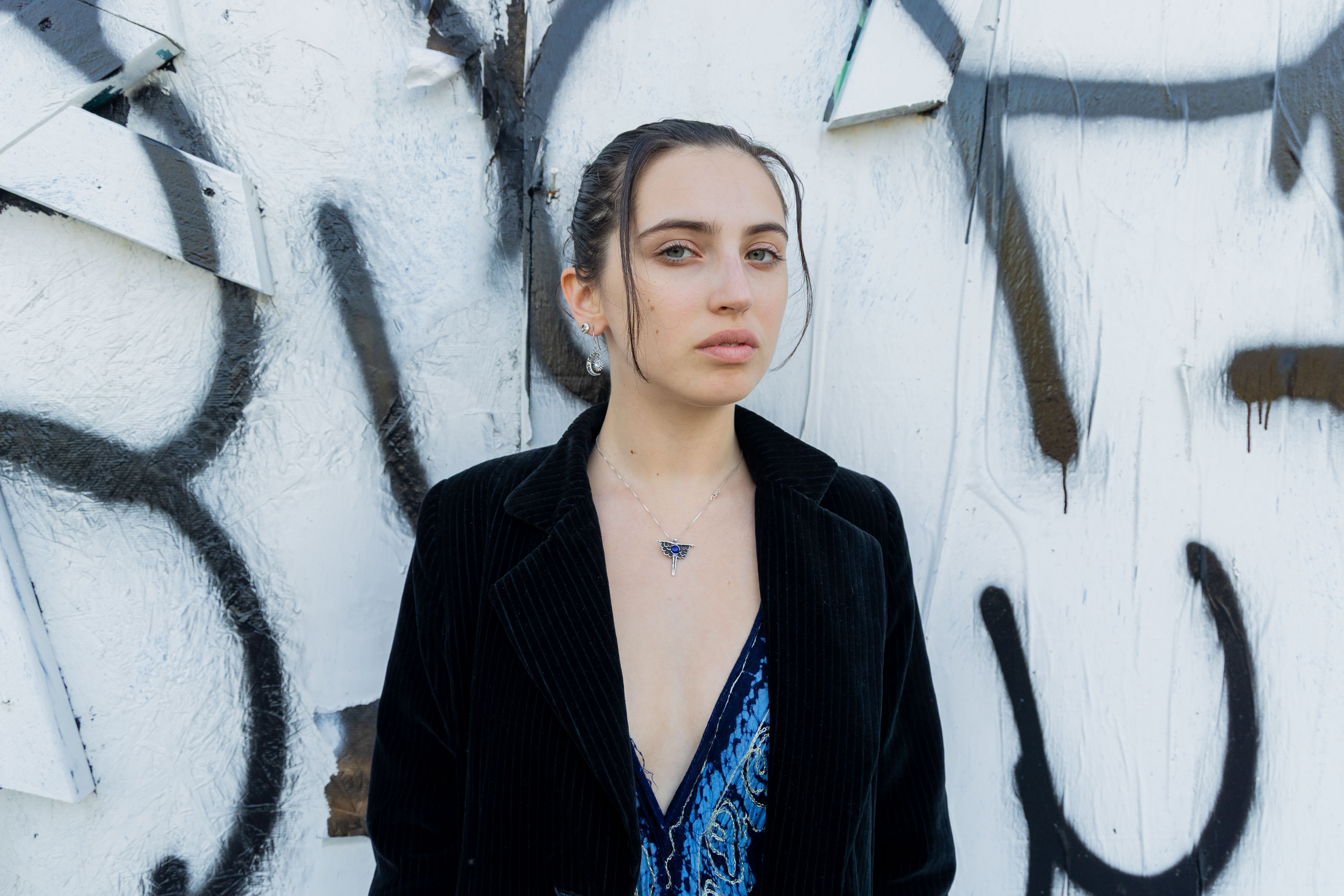 Dry stands in front of a white wall with black graffiti. Throughout her journey as a music artist, the 2024 graduate said she has had the opportunity to learn more about herself, attempting to capture incomprehensible feelings into her work. (Chenrui Zhang/Daily Bruin)