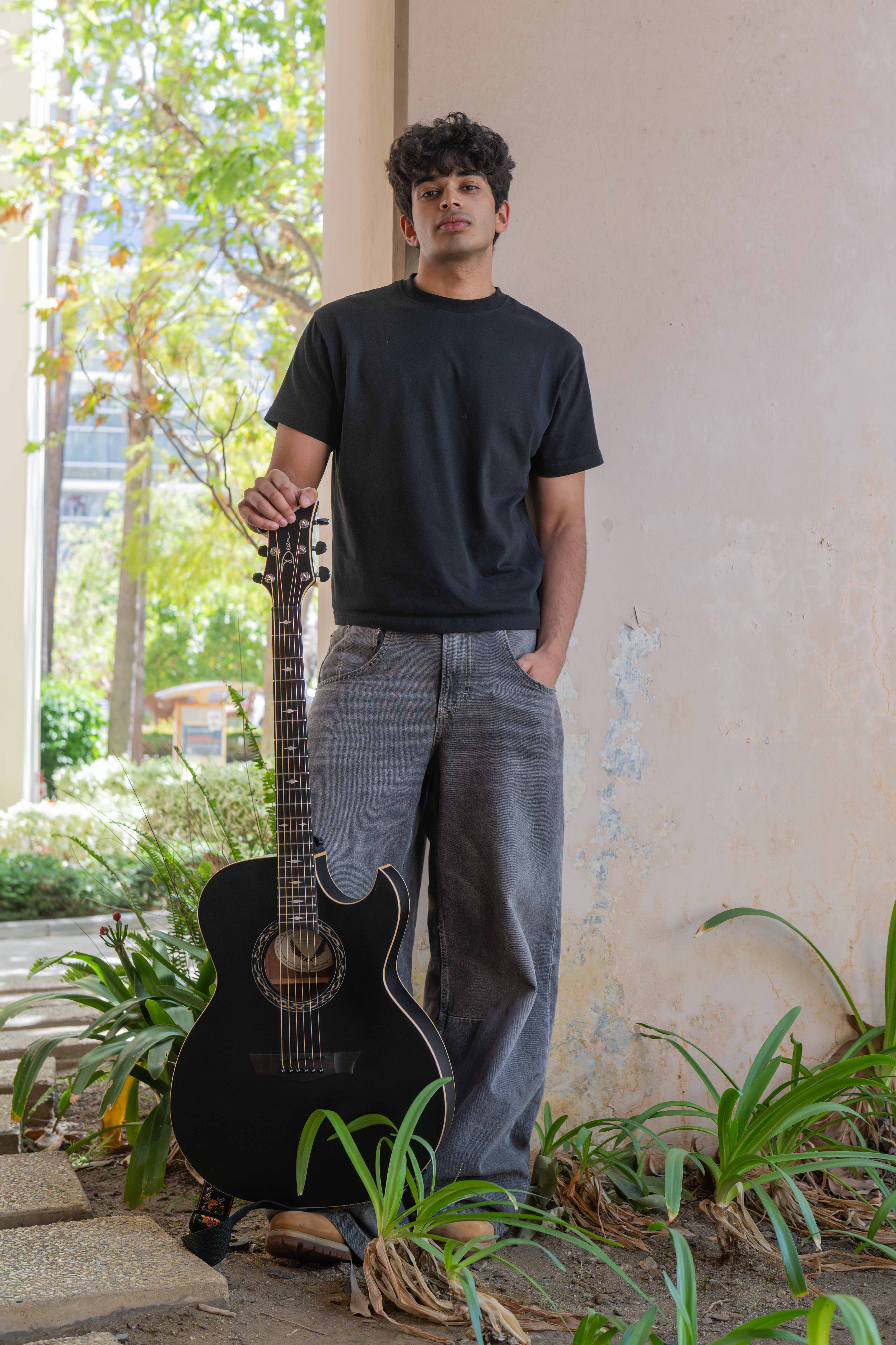 Aror is standing against a wall wearing a black shirt and gray pants, with his guitar propped on his boot. The music industry student described his sound as folk and indie-pop, taking inspiration from artists like Adrianne Lenker and Lizzy McAlpine. (Leydi Cris Cobo Cordon/Daily Bruin senior staff)