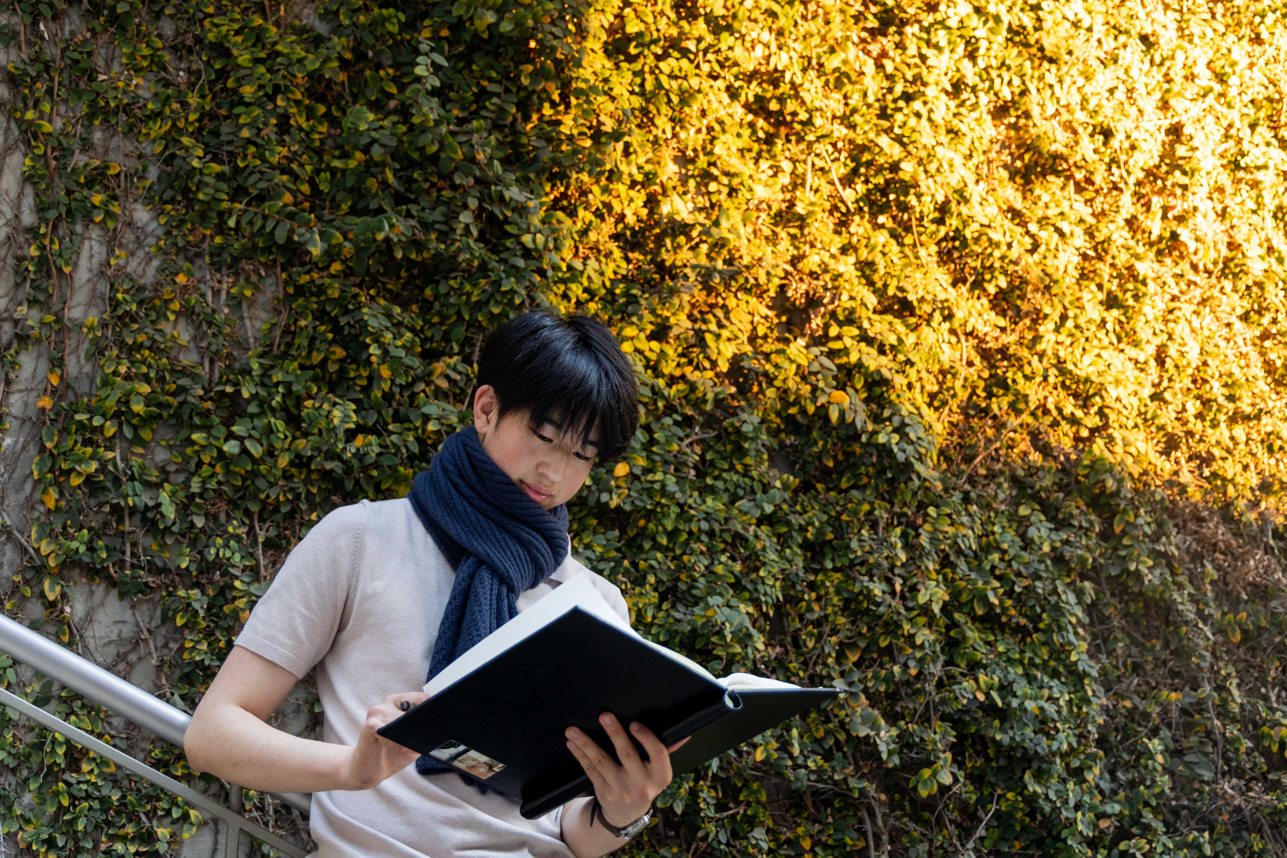 Su stands on a staircase in front of a wall of greenery, wearing a blue scarf as he flips through a notebook. (Chenrui Zhang/Daily Bruin staff)