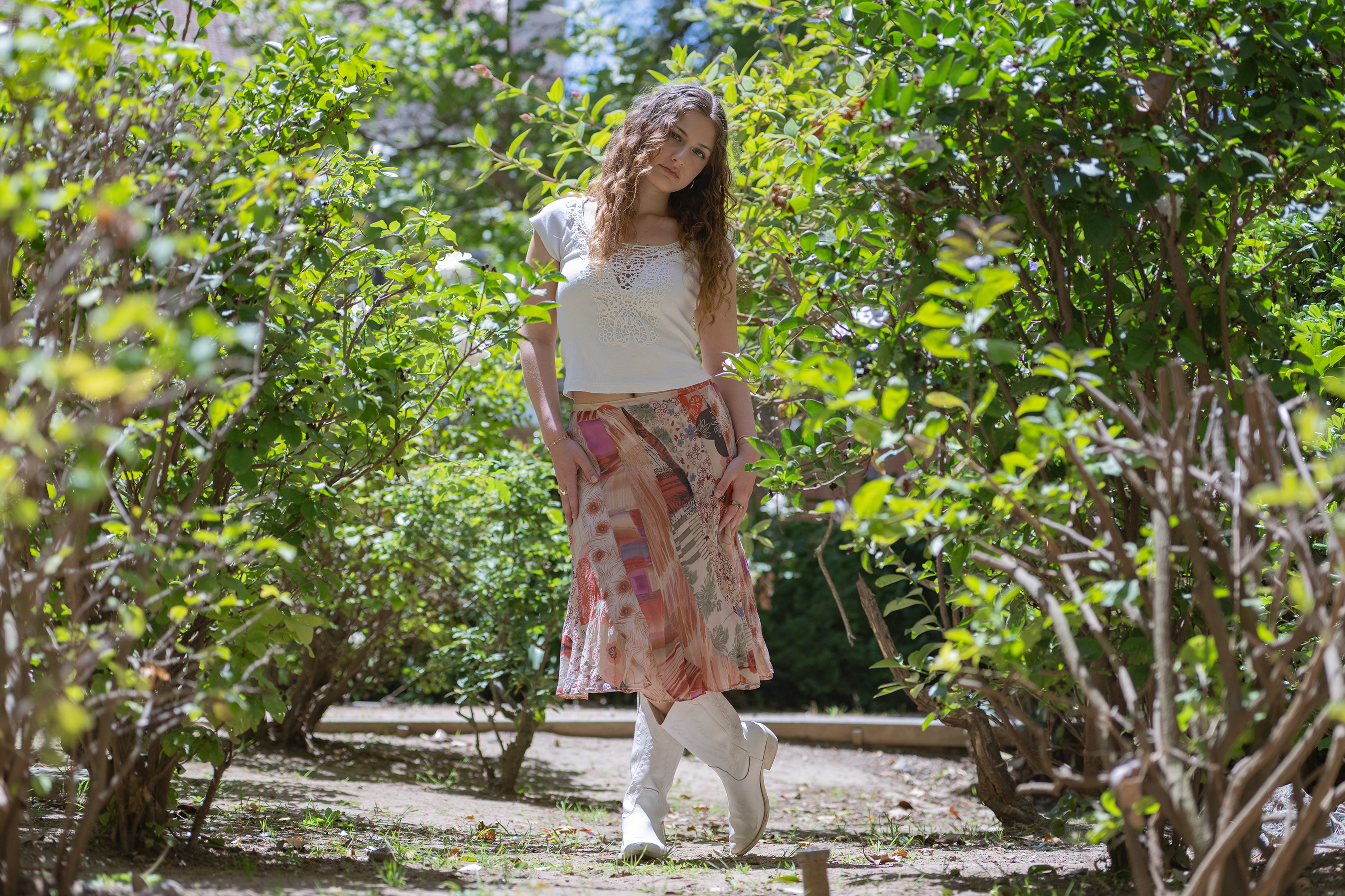 Wertheimer stands in a path between trees, wearing a long multi-pattern skirt and white boots. As a music industry and musicology minor, she said she developed her confidence in performing through classes at the UCLA Herb Alpert School of Music. (Presley Liu/Daily Bruin)