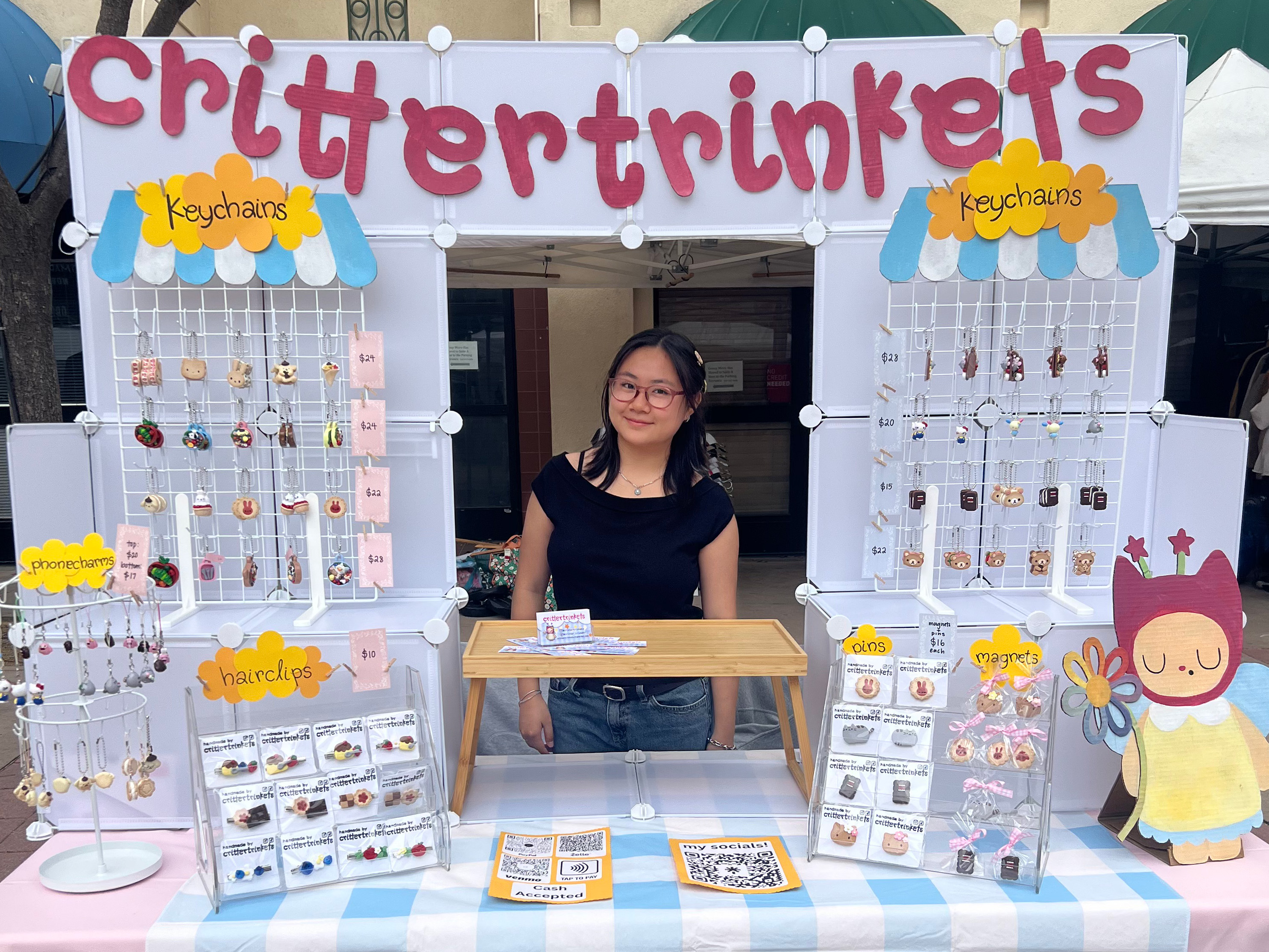Donna Wu poses behind a vendor table full of her merchandise, or trinkets, at the Bruin Flea. Wu launched Critter Trinkets in October 2025 and makes all her clay pieces by hand. (Courtesy of Donna Wu. Cropped from original)